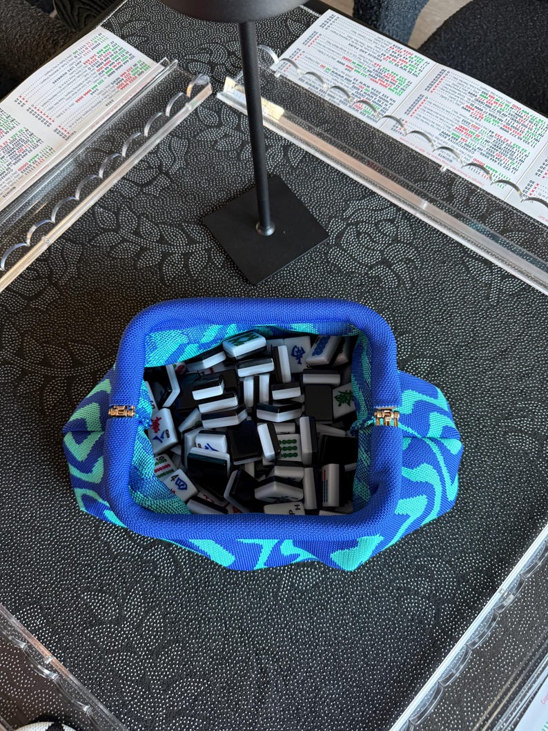 May include: A blue and teal patterned fabric pouch with a metal frame closure, filled with a collection of small, rectangular black and white Mahjong tiles featuring various engraved symbols and characters in green, red, and blue ink.