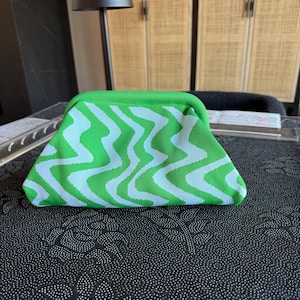 May include: A clutch bag featuring a vibrant green and white wavy, zigzag pattern. The bag has a structured, curved top frame closure and a soft, knit-like texture. The design consists of repeating vertical undulating lines in bright green.