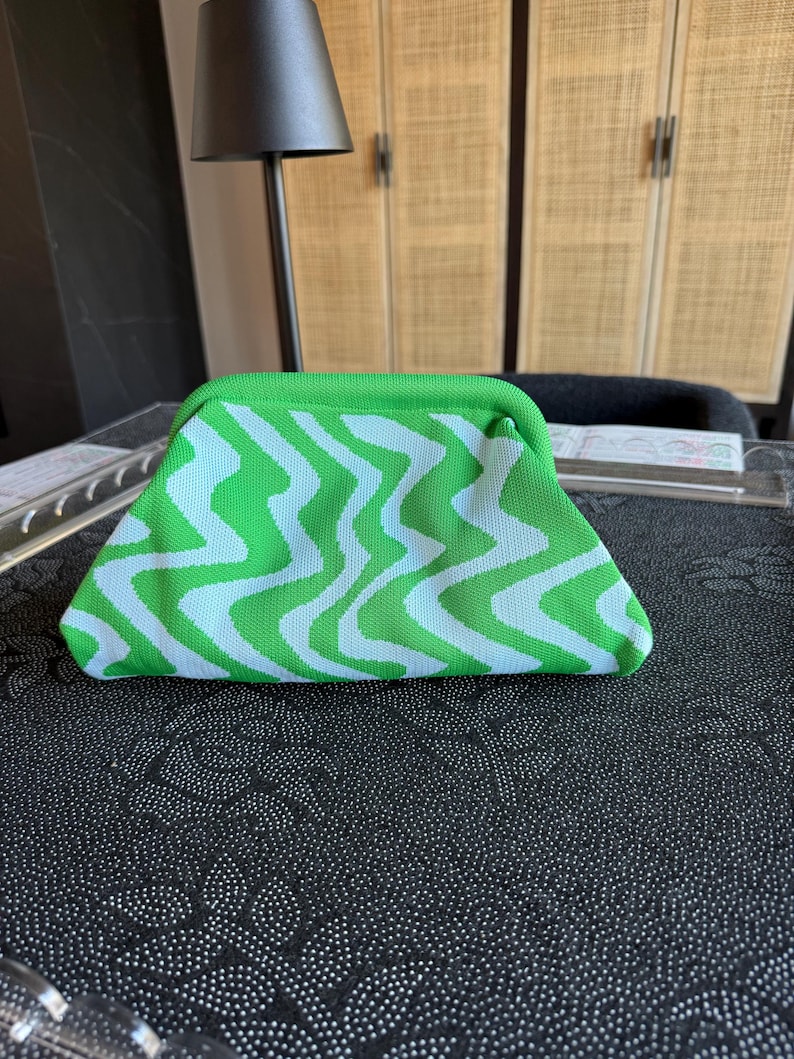 May include: A clutch bag featuring a vibrant green and white wavy, zigzag pattern. The bag has a structured, curved top frame closure and a soft, knit-like texture. The design consists of repeating vertical undulating lines in bright green.