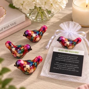 May include: Three small bird-shaped figurines made of clear resin filled with colorful beads and gold flakes, alongside a white organza drawstring bag containing a fourth bird figurine and a card titled OH MY TINY RACK BIRD with text.