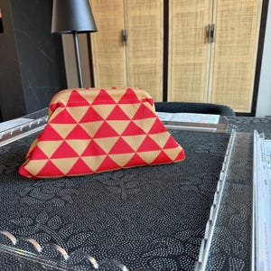 May include: A structured pouch featuring a repeating geometric pattern of red and tan triangles. The bag has a curved top frame closure and a fabric exterior with a visible woven texture, presenting a triangular silhouette when viewed from the front.