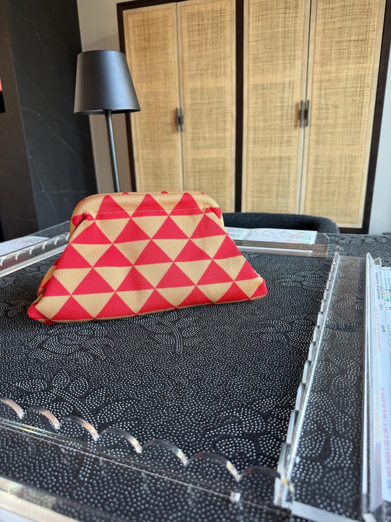 May include: A structured pouch featuring a repeating geometric pattern of red and tan triangles. The bag has a curved top frame closure and a fabric exterior with a visible woven texture, presenting a triangular silhouette when viewed from the front.