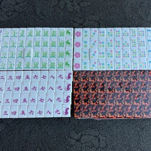 K&ouml;nnte beinhalten: Ein Set rechteckiger Mahjong-Spielsteine mit wei&szlig;en Vorderseiten, die gr&uuml;ne Bambusmotive, rosa Blumen, blaue Symbole und rote chinesische Schriftzeichen zeigen. Ein Teil der Steine ist in Klarsichtfolie verpackt und hat ein braun-schwarzes Schildpattmuster.