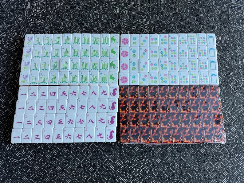K&ouml;nnte beinhalten: Ein Set rechteckiger Mahjong-Spielsteine mit wei&szlig;en Vorderseiten, die gr&uuml;ne Bambusmotive, rosa Blumen, blaue Symbole und rote chinesische Schriftzeichen zeigen. Ein Teil der Steine ist in Klarsichtfolie verpackt und hat ein braun-schwarzes Schildpattmuster.