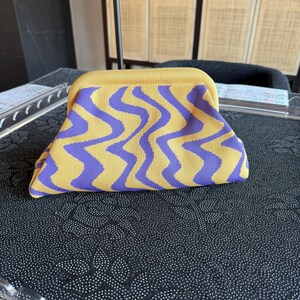 May include: A structured clutch bag featuring a vibrant yellow and purple wavy, zig-zag pattern. The bag has a curved top frame closure and a soft, knit-like texture, presenting a distinct geometric design across the entire exterior surface.
