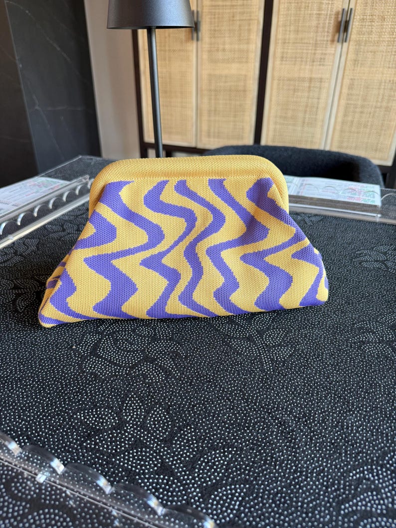 May include: A structured clutch bag featuring a vibrant yellow and purple wavy, zig-zag pattern. The bag has a curved top frame closure and a soft, knit-like texture, presenting a distinct geometric design across the entire exterior surface.