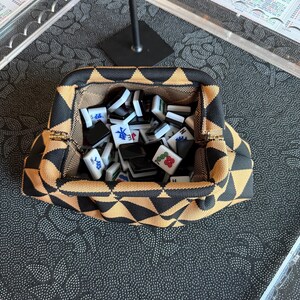 May include: A structured fabric pouch featuring a black and tan geometric triangle pattern, containing a collection of white and black mahjong tiles with various red, blue, and green character markings and symbols printed on their surfaces.