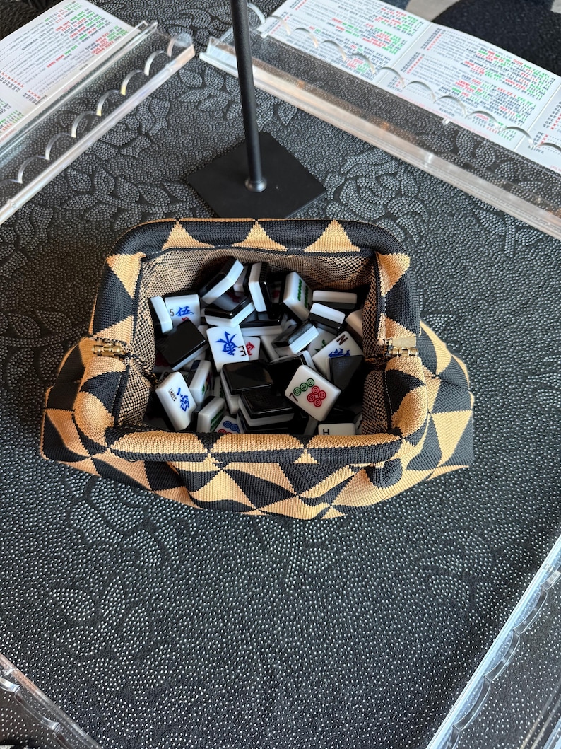 May include: A structured fabric pouch featuring a black and tan geometric triangle pattern, containing a collection of white and black mahjong tiles with various red, blue, and green character markings and symbols printed on their surfaces.