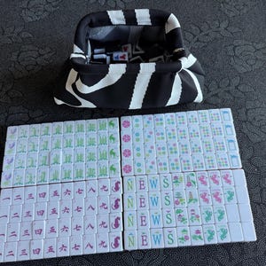 K&ouml;nnte beinhalten: Ein schwarz-wei&szlig; gestreifter Stoffbeutel mit Metallrahmenverschluss, der wei&szlig;e Mahjong-Steine enth&auml;lt. Unter dem Beutel liegen Reihen wei&szlig;er Mahjong-Steine mit gr&uuml;nen Bambusmotiven, bunten Blumenmustern, chinesischen Schriftzeichen sowie den Buchstaben N, E, W, S.