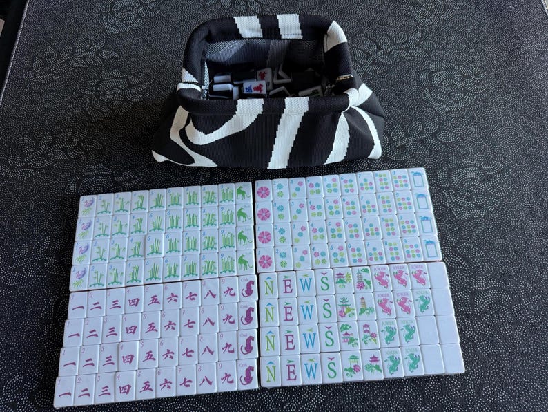 K&ouml;nnte beinhalten: Ein schwarz-wei&szlig; gestreifter Stoffbeutel mit Metallrahmenverschluss, der wei&szlig;e Mahjong-Steine enth&auml;lt. Unter dem Beutel liegen Reihen wei&szlig;er Mahjong-Steine mit gr&uuml;nen Bambusmotiven, bunten Blumenmustern, chinesischen Schriftzeichen sowie den Buchstaben N, E, W, S.