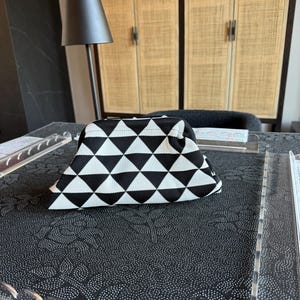 May include: A structured pouch featuring a repeating black and white geometric triangle pattern. The bag has a curved top frame closure and a textured fabric exterior, presenting a consistent monochromatic design across the entire surface of the item.