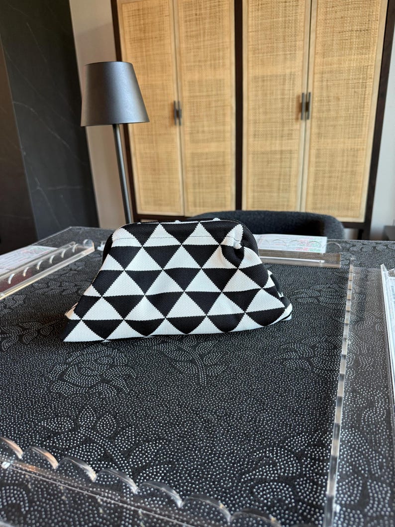 May include: A structured pouch featuring a repeating black and white geometric triangle pattern. The bag has a curved top frame closure and a textured fabric exterior, presenting a consistent monochromatic design across the entire surface of the item.