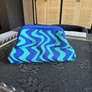 May include: A structured clutch bag featuring a vibrant wavy pattern in alternating shades of royal blue and bright turquoise. The bag has a curved top frame closure and a textured, knit-like fabric construction with a solid blue top border.