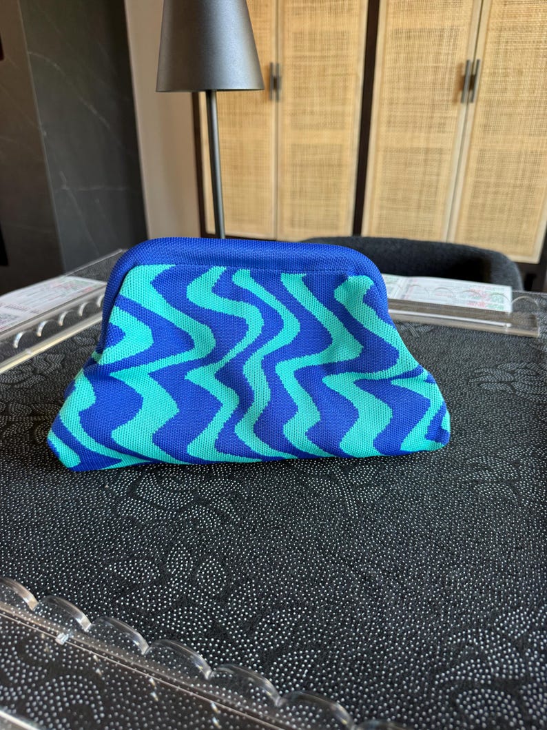 May include: A structured clutch bag featuring a vibrant wavy pattern in alternating shades of royal blue and bright turquoise. The bag has a curved top frame closure and a textured, knit-like fabric construction with a solid blue top border.