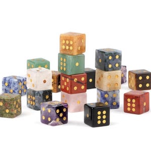May include: A collection of gemstone dice in various colors, including green, red, blue, and brown. Each die features gold-colored dots representing the numbers. The dice are stacked and arranged, showcasing their unique textures and hues.
