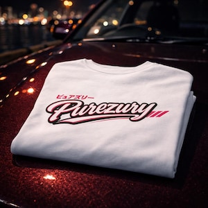 May include: A folded white t-shirt with the word "Purezury" in a stylized script, outlined in black and accented with pink. Japanese characters are above the word. The shirt is resting on a red car hood.