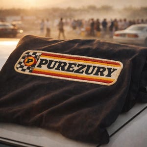 May include: A dark gray fabric with a rectangular patch featuring the word "PUREZURY" in a bold, retro font. The patch has a checkered flag design and a red, yellow, and orange stripe.