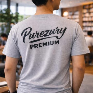 May include: A gray t-shirt with the text "Purezury PREMIUM" printed on the back in black. The shirt is short-sleeved and appears to be made of a soft material. The background is blurred, suggesting an indoor setting.