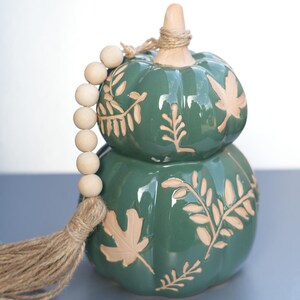 Könnte beinhalten: Ein zweistöckiger, grüner Keramik-Kürbis mit Blatt- und Rankenmustern. Eine Kette aus Holzperlen und eine Quaste aus Jute sind befestigt. Der Kürbis ist ein dekorativer Artikel.