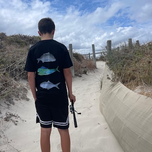 Puede incluir: Una camiseta negra con tres ilustraciones de peces en la espalda, combinada con pantalones cortos negros con rayas blancas. La persona camina por un sendero arenoso, sosteniendo una caña de pescar, bajo un cielo nublado.