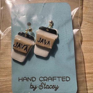 May include: A pair of handmade coffee cup earrings. Each earring is a miniature replica of a coffee cup, featuring a black lid, white cup, and a brown band that reads "JAVA". The earrings are attached to gold-colored posts.