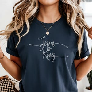 May include: A dark navy blue short-sleeve t-shirt featuring the white cursive text "Jesus is King" centered on the chest. The shirt has a crew neckline and is paired with blue denim shorts. The design is printed in a thin, elegant script font.