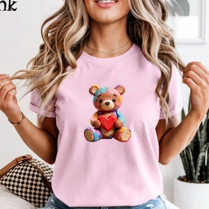 Peut inclure: T-shirt rose clair avec un ours en peluche coloré tenant un cœur rouge. L'ours en peluche a un design patchwork avec différentes couleurs. Le mot "Pink" est dans le coin supérieur gauche de l'image.