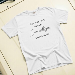 Puede incluir: Camiseta blanca con el texto "You are not alone. I am with you. Isaiah 41:10" en escritura negra. La camiseta es de material suave y tiene cuello redondo.