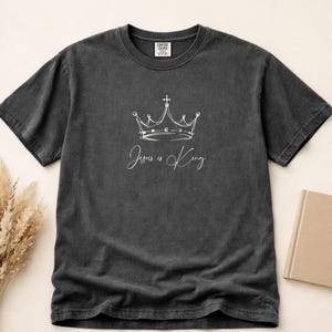 Könnte beinhalten: Dunkelgraues T-Shirt mit weißem Kronen-Design und dem Text "Jesus is King". Das Shirt hat kurze Ärmel und einen Rundhalsausschnitt. Das Design befindet sich mittig auf der Vorderseite.