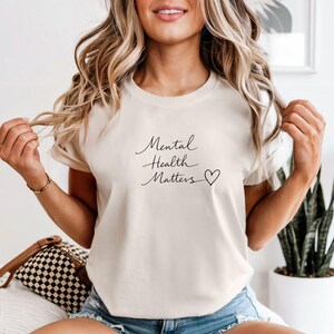 May include: Cream-colored t-shirt with the handwritten text "Mental Health Matters" and a heart symbol. The shirt is a short-sleeved crew neck style.