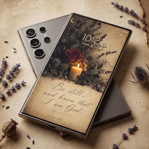 May include: A silver smartphone with a screen displaying a floral design, a lit candle, and the text "Be still, and know that I am God." The time on the screen reads 10:22. The phone is resting on a textured, light brown surface.