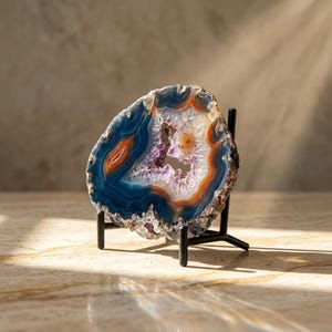 May include: A striking geode slice, displaying bands of blue, orange, and white, with a crystalline interior of purple and white. The polished stone is presented on a black metal stand, against a neutral backdrop.