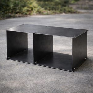 May include: A dark gray metal coffee table with two open compartments. The table is rectangular with a flat top and is made of folded metal sheets. The table is sitting on a gray concrete surface.
