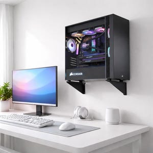 Puede incluir: Una caja de ordenador negra montada en una pared blanca, con un panel lateral transparente que revela los componentes internos. La caja está soportada por soportes negros. Un monitor de ordenador, teclado, ratón y otros accesorios están sobre un escritorio blanco.