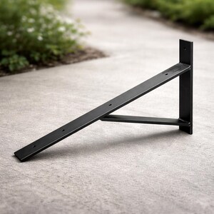 May include: A black metal shelf bracket with a right-angle design. The bracket features a flat, horizontal support arm with pre-drilled holes for mounting, and a vertical support piece. The bracket is designed to support a shelf and is suitable for various home decor styles.