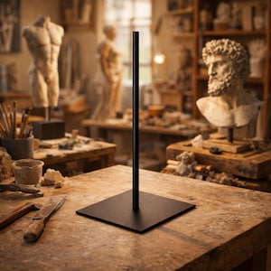 Puede incluir: Un soporte de exhibición de metal negro con una base cuadrada y un poste vertical. El soporte está sobre una mesa de madera en el estudio de un artista, con esculturas y materiales de arte en el fondo.