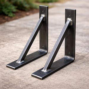 May include: Two industrial-style metal shelf brackets. Each bracket has a rectangular base and a vertical support, connected by a diagonal brace. The brackets are made of dark gray metal with visible weld marks and mounting holes.