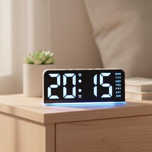 May include: A white digital alarm clock showing the time 20:15 with blue LED lighting. The clock also displays the days of the week. A small succulent plant in a white pot is to the left of the clock on a wooden bedside table.