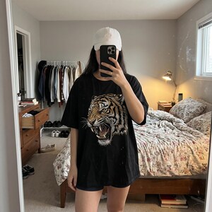 May include: A black short-sleeved t-shirt featuring a large, detailed graphic print of a roaring tiger head in shades of gold, orange, and white. The shirt is paired with a plain white baseball cap, creating a casual look for everyday wear.