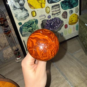 May include: A spherical object with a polished, glossy surface featuring swirling patterns of deep orange, rust red, and brown. The sphere is attached to a cylindrical handle, creating a knob-like structure with a smooth, reflective finish and distinct color variations throughout the stone-like material.