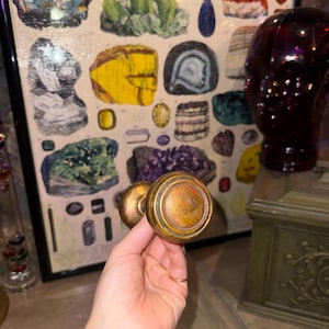 May include: A round, metallic door knob with a weathered, tarnished brass finish. The knob features a circular, stepped design with concentric rings and a slightly convex center, showing signs of age and oxidation across its surface.
