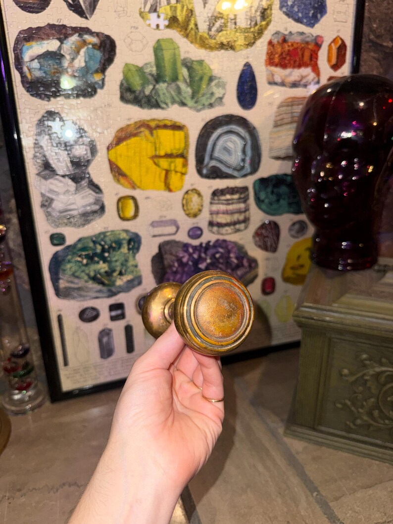 May include: A round, metallic door knob with a weathered, tarnished brass finish. The knob features a circular, stepped design with concentric rings and a slightly convex center, showing signs of age and oxidation across its surface.