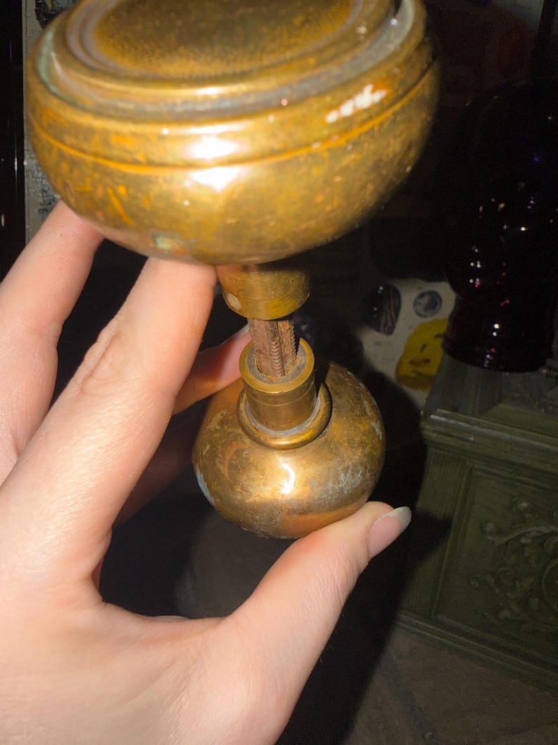 May include: A vintage brass door knob set featuring two spherical knobs connected by a central threaded metal spindle. The brass surface shows signs of age, including a dull patina and minor surface wear, with a smooth, rounded shape for each knob.