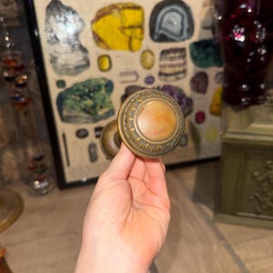 May include: A round, antique-style brass door knob featuring a smooth, flat center disc surrounded by a decorative, raised border with a repeating scalloped pattern. The metal surface displays a warm, aged patina with visible signs of wear.