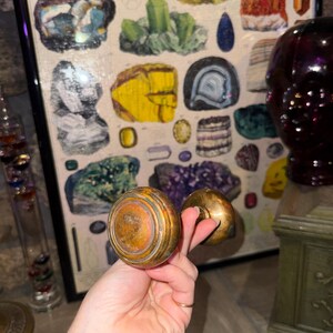 May include: A pair of round, metallic door knobs with a weathered, tarnished brass finish. The knobs feature a concentric circular design on the face and are connected by a central spindle, showcasing a vintage appearance with visible surface wear.