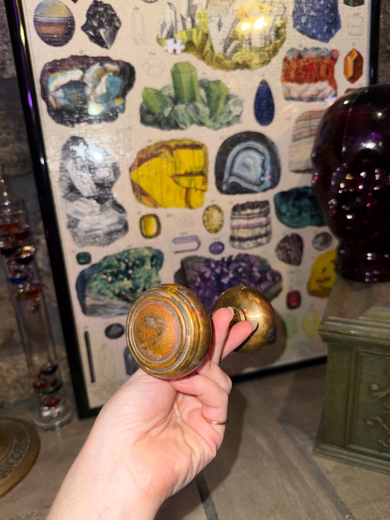May include: A pair of round, metallic door knobs with a weathered, tarnished brass finish. The knobs feature a concentric circular design on the face and are connected by a central spindle, showcasing a vintage appearance with visible surface wear.