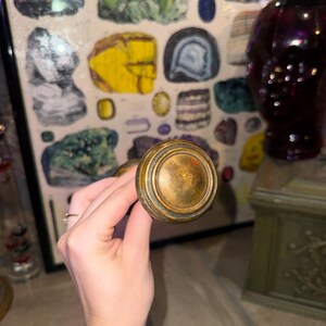 May include: A round, metallic door knob with a weathered, antique brass finish. The knob features a circular, stepped design with a smooth, slightly domed center and concentric ridges around the edge, showing signs of age and patina.