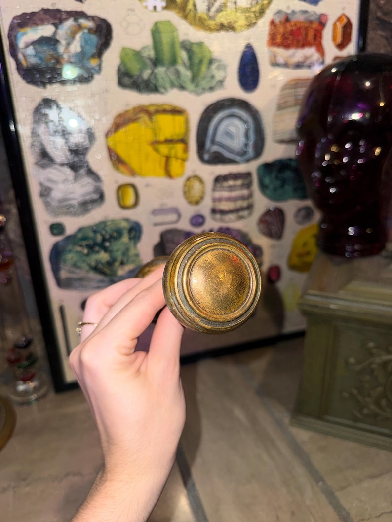 May include: A round, metallic door knob with a weathered, antique brass finish. The knob features a circular, stepped design with a smooth, slightly domed center and concentric ridges around the edge, showing signs of age and patina.