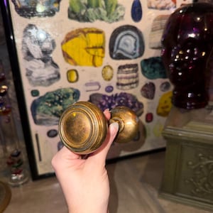 May include: A round, metallic door knob with a weathered, antique brass finish. The knob features a circular, stepped design on its face and a cylindrical neck connecting to a smaller, matching knob component, showing a tarnished patina.