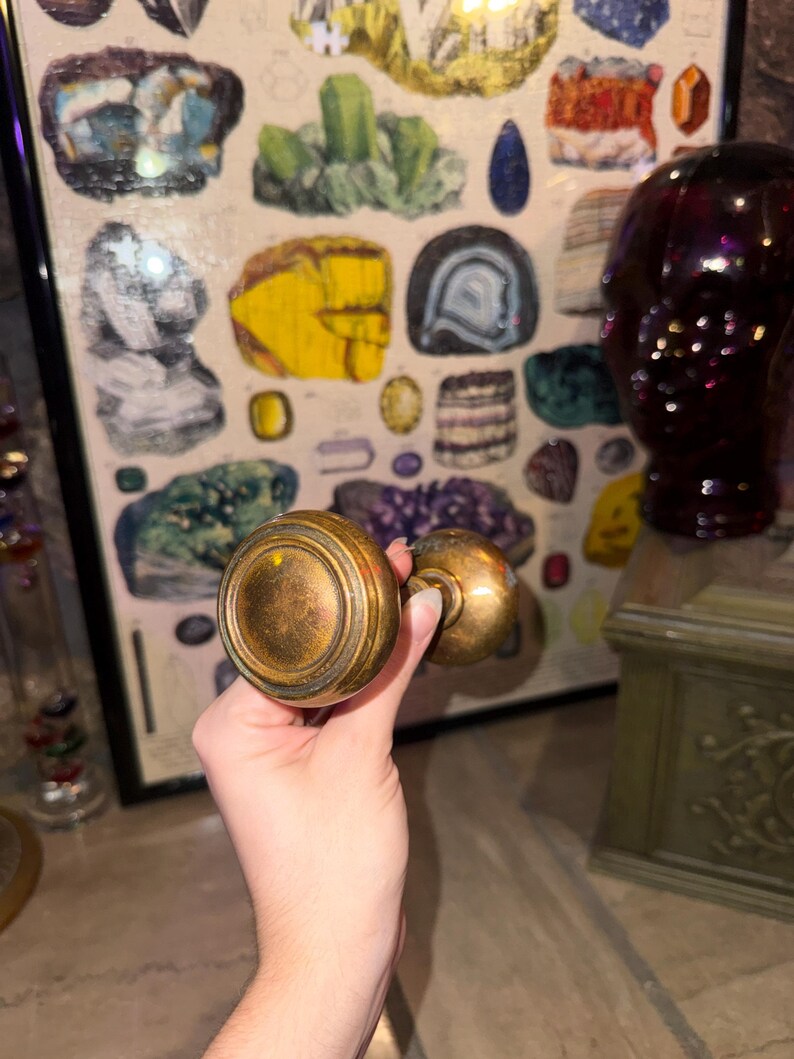 May include: A round, metallic door knob with a weathered, antique brass finish. The knob features a circular, stepped design on its face and a cylindrical neck connecting to a smaller, matching knob component, showing a tarnished patina.